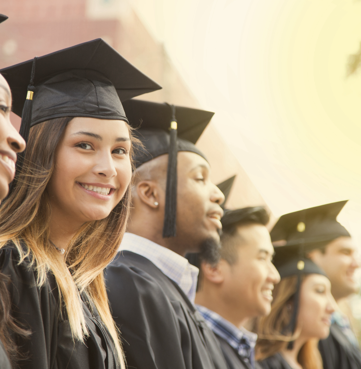 Latin descent girl waits in line during college graduation ceremony.   She looks at camera with a big smile as she wears a black cap and gown.