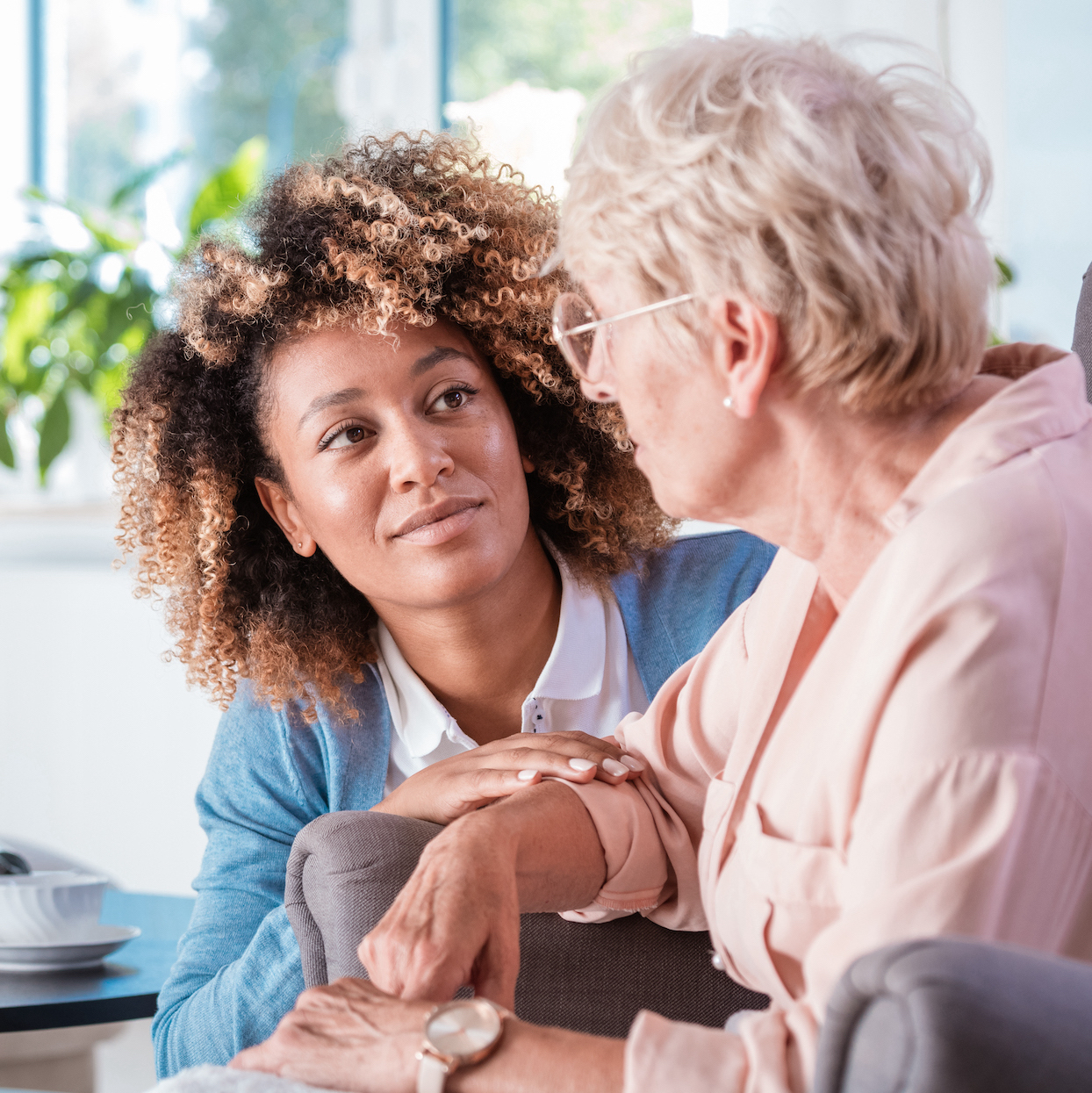 Female home caregiver talking with senior woman, sitting in living room and listening to her carefully.