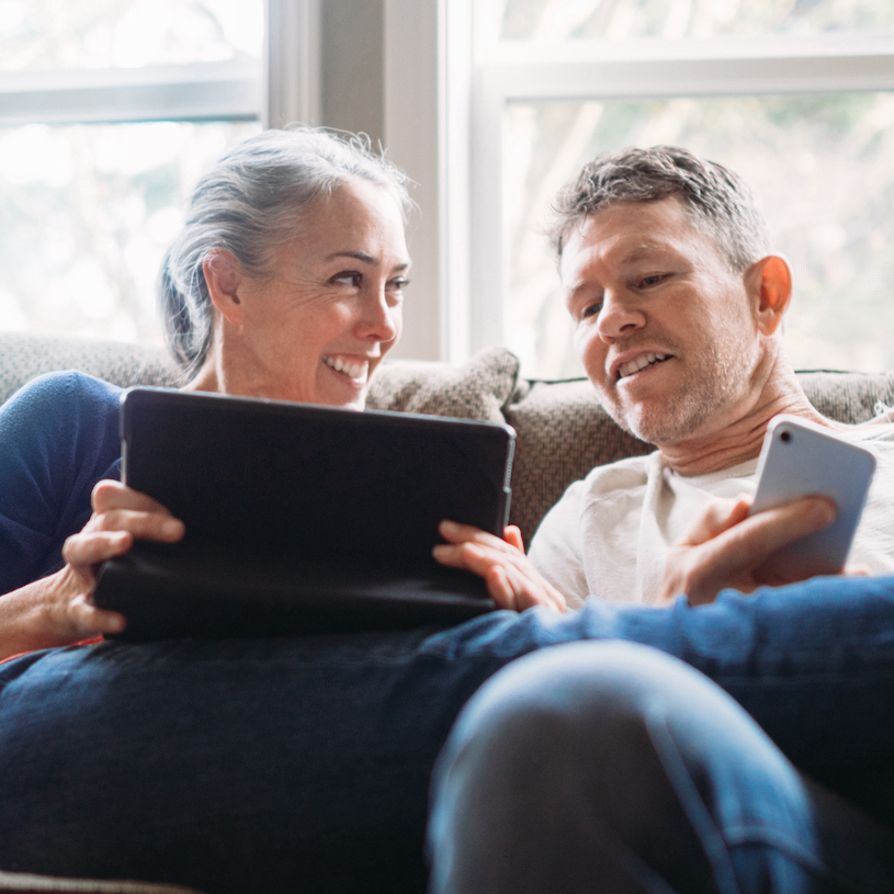 A couple in their 50's relax in their home on the living room couch, enjoying reading and surfing the internet on their mobile touchscreen phones and computer tablet.